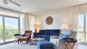A spacious living room with a large window, a ceiling fan, a blue sofa, a wooden coffee table, and a wicker chair.