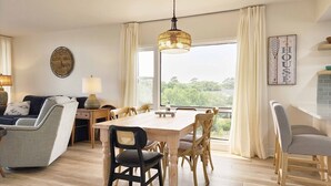 A living room with a dining table, chairs, a sofa, a lamp, a clock, and a view of greenery outside.