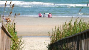 A beach with a wooden boardwalk leading to a sandy shore. There are three beach chairs and two people sitting in them.