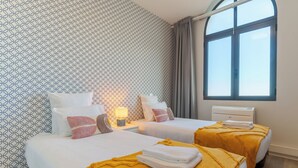 A hotel room with two beds, a patterned wall, a large window, and a bedside table with a lamp. - Casa Bela Vista (São Gonçalo)