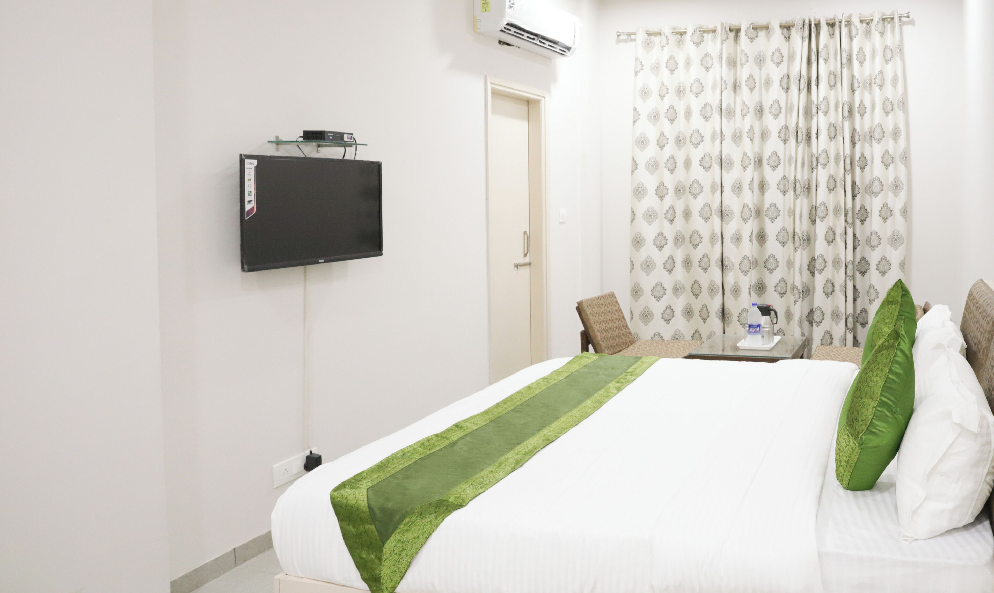 Una habitación de hotel con cama, televisor montado en la pared, cafetera en la mesita de noche y cortina estampada.