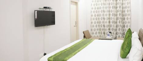 A hotel room with a bed, a TV mounted on the wall, a coffee maker on the nightstand, and a patterned curtain.