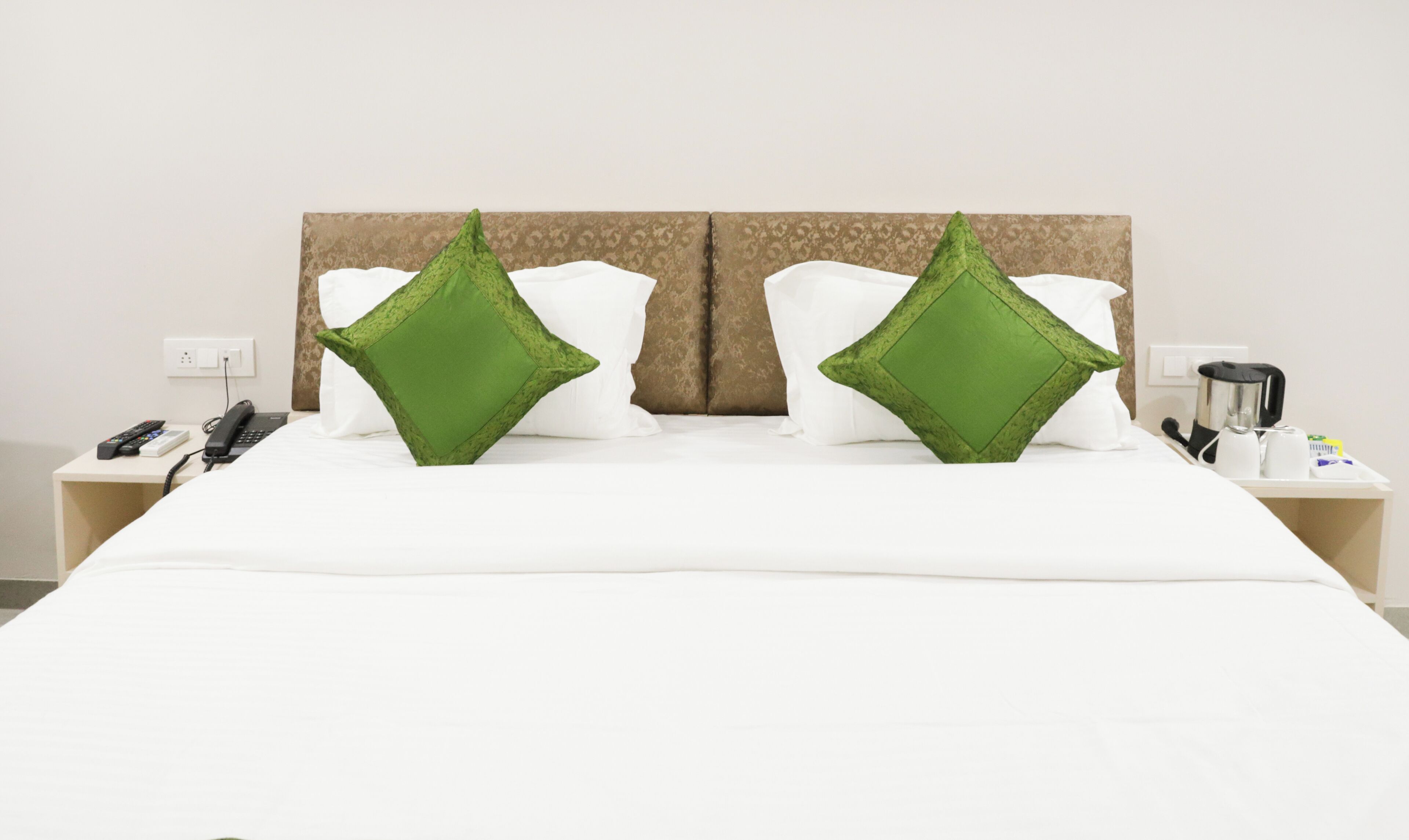 A hotel room with a bed, two green pillows, a bedside table with a phone, a remote, and a coffee maker.