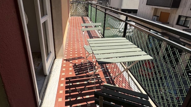 A balcony with a red patterned floor, a green metal railing, and a white building in the background.