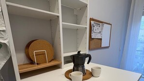 A white kitchen counter with a coffee maker, two white cups, and a cork coaster. There is a cork mat on the counter and a cork coaster on the shelf.
