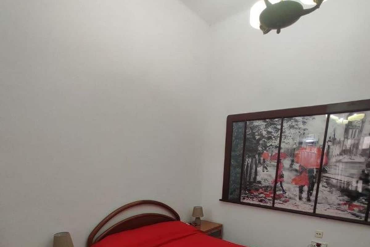 A bedroom with a single bed, a nightstand with a lamp, a wall-mounted mirror, and a ceiling light fixture.
