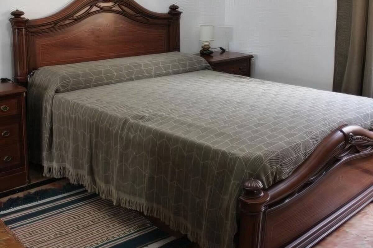 A single bed with a wooden headboard and footboard, a bedside table, and a rug on a tiled floor.