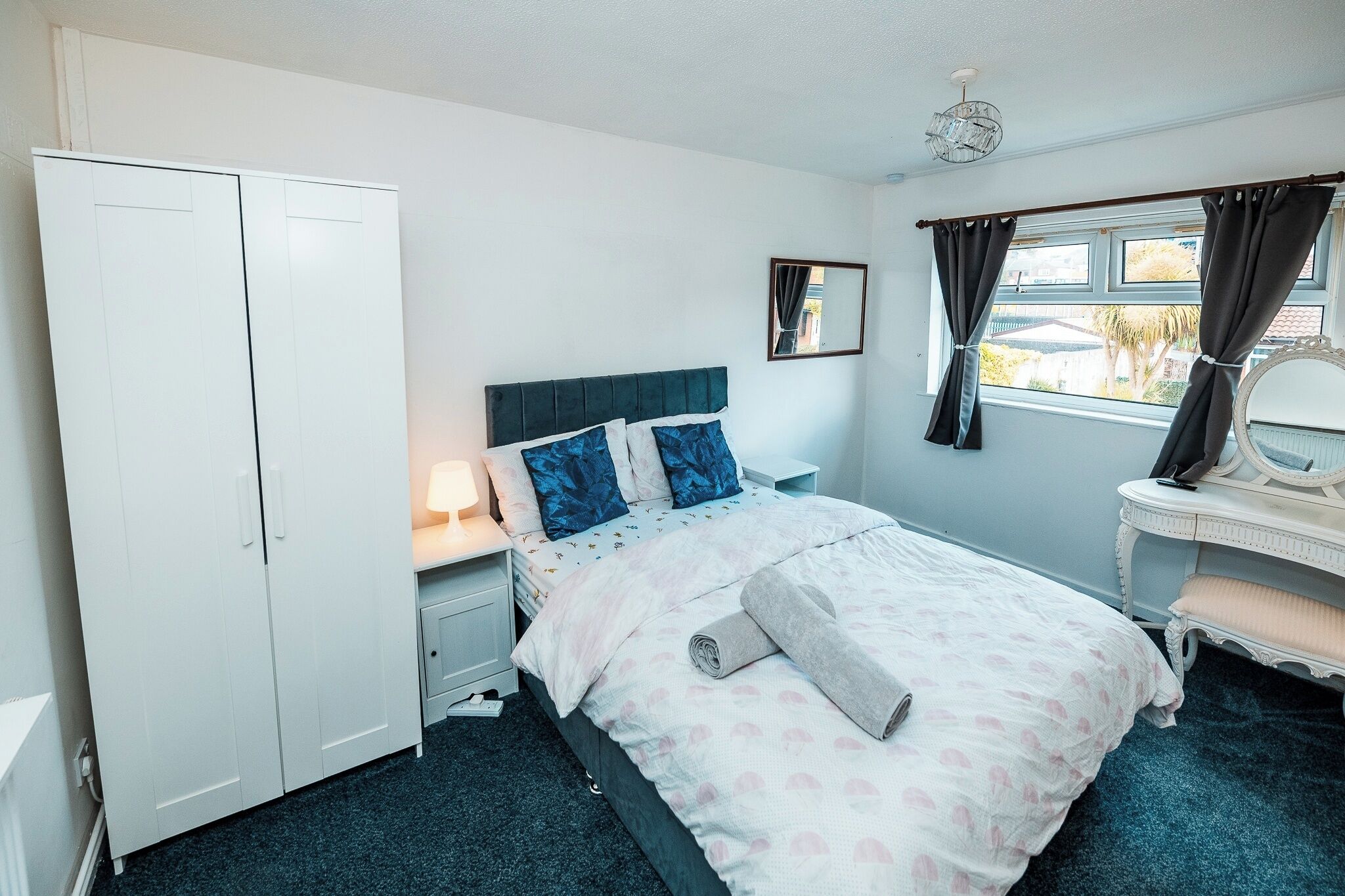 A bedroom with a bed, bedside table, lamp, wardrobe, window with curtains, and a mirror.
