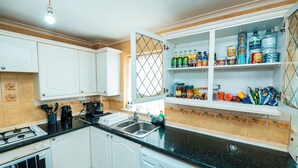 Een keuken met witte kasten, een zwart aanrecht, een gasfornuis, een wasmachine en planken vol drankjes en snacks.