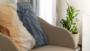 En beige soffa med en mångfärgad kudde, en krukväxt och ett fönster med gardiner.