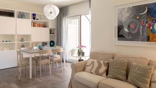 Ett modernt vardagsrum med en beige soffa, ett matbord med stolar och en bokhylla.