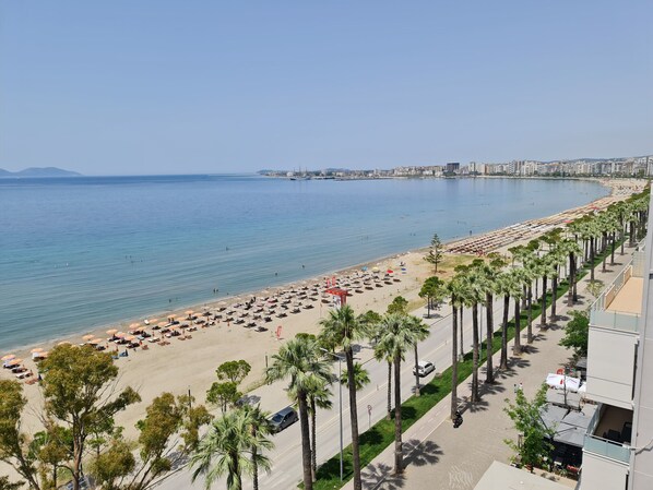 A coastal hotel with a beach view, a promenade lined with palm trees, and a cityscape in the background.