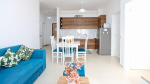 A modern living room with a blue sofa, white dining table, and chairs. A kitchen area is visible in the background with wooden cabinets and a refrigerator.