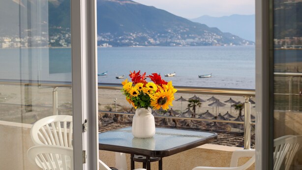 A balcony with a table and chairs, a vase of sunflowers, and a view of the sea and mountains.