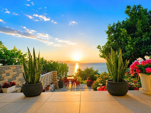 A patio with potted plants, a table, and chairs, overlooking a sunset over the sea.