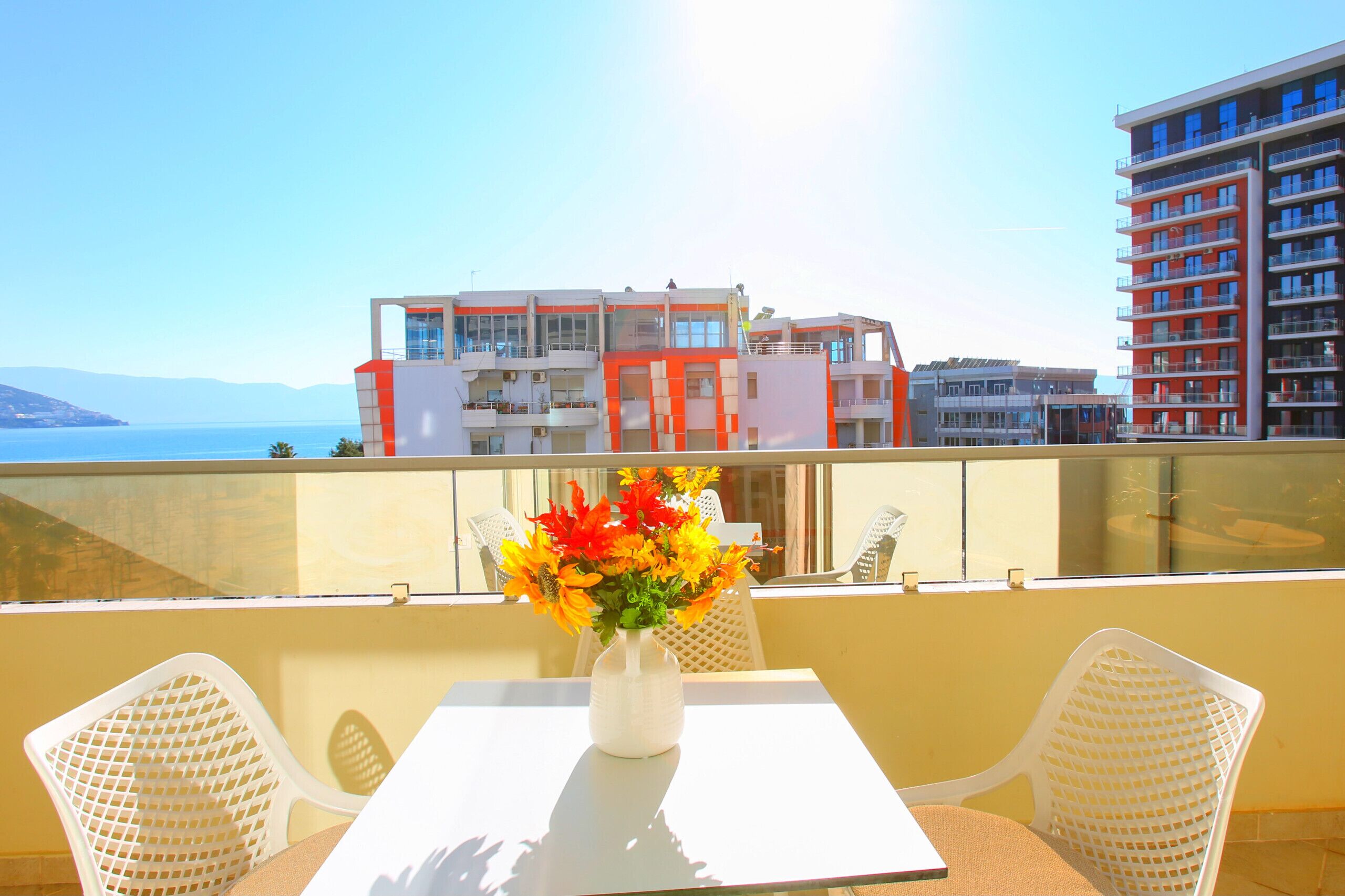 Ein Balkon mit Tisch und Stühlen, einem Blumenstrauß und Blick auf ein Gebäude mit Balkonen sowie ein Gewässer im Hintergrund.