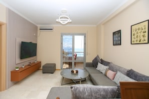 A modern living room with a flat-screen TV, a grey sofa, a coffee table, and a balcony with a view.