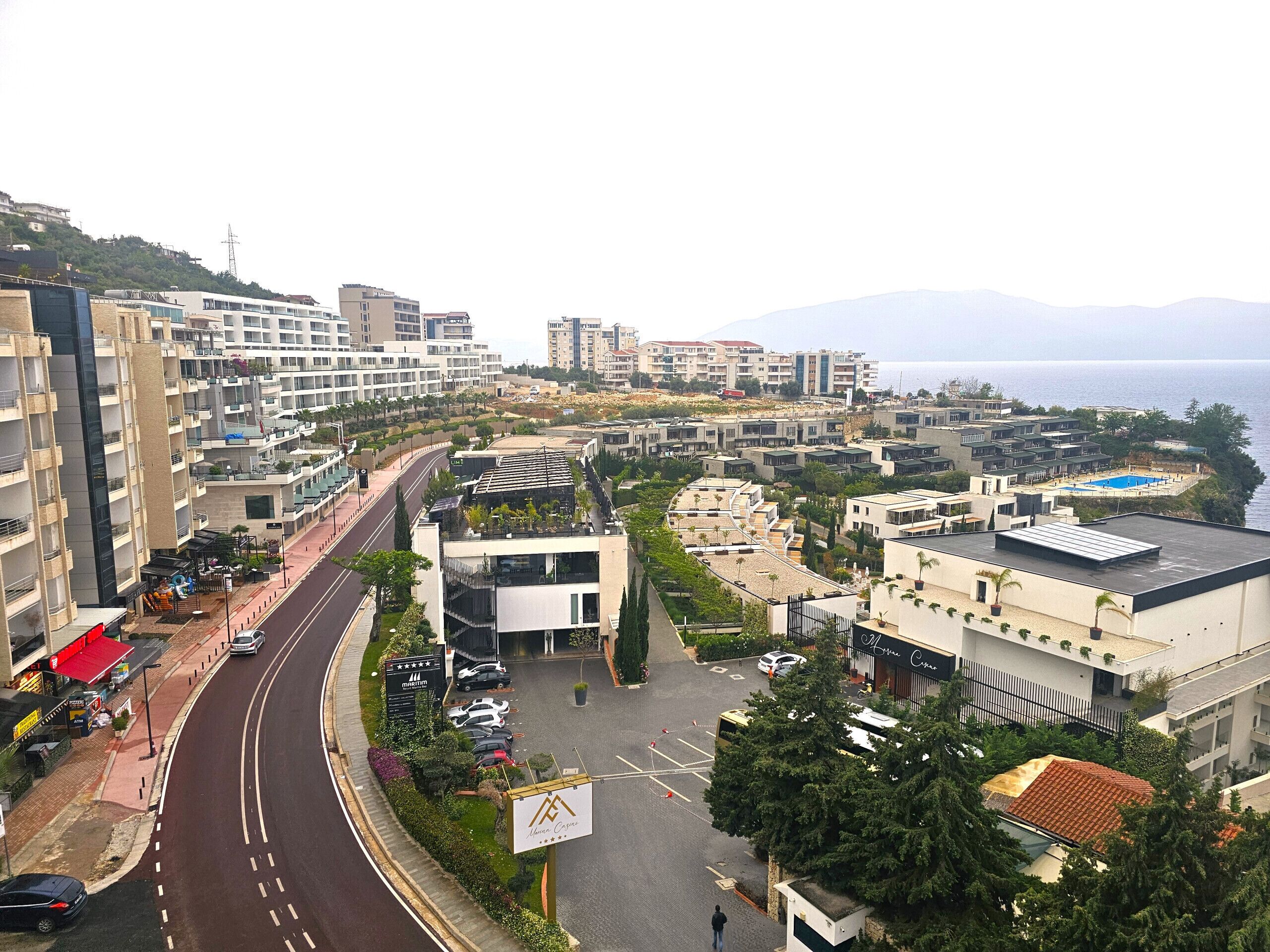 Zona urbana moderna con edificios residenciales y comerciales, una carretera sinuosa y vista al mar.