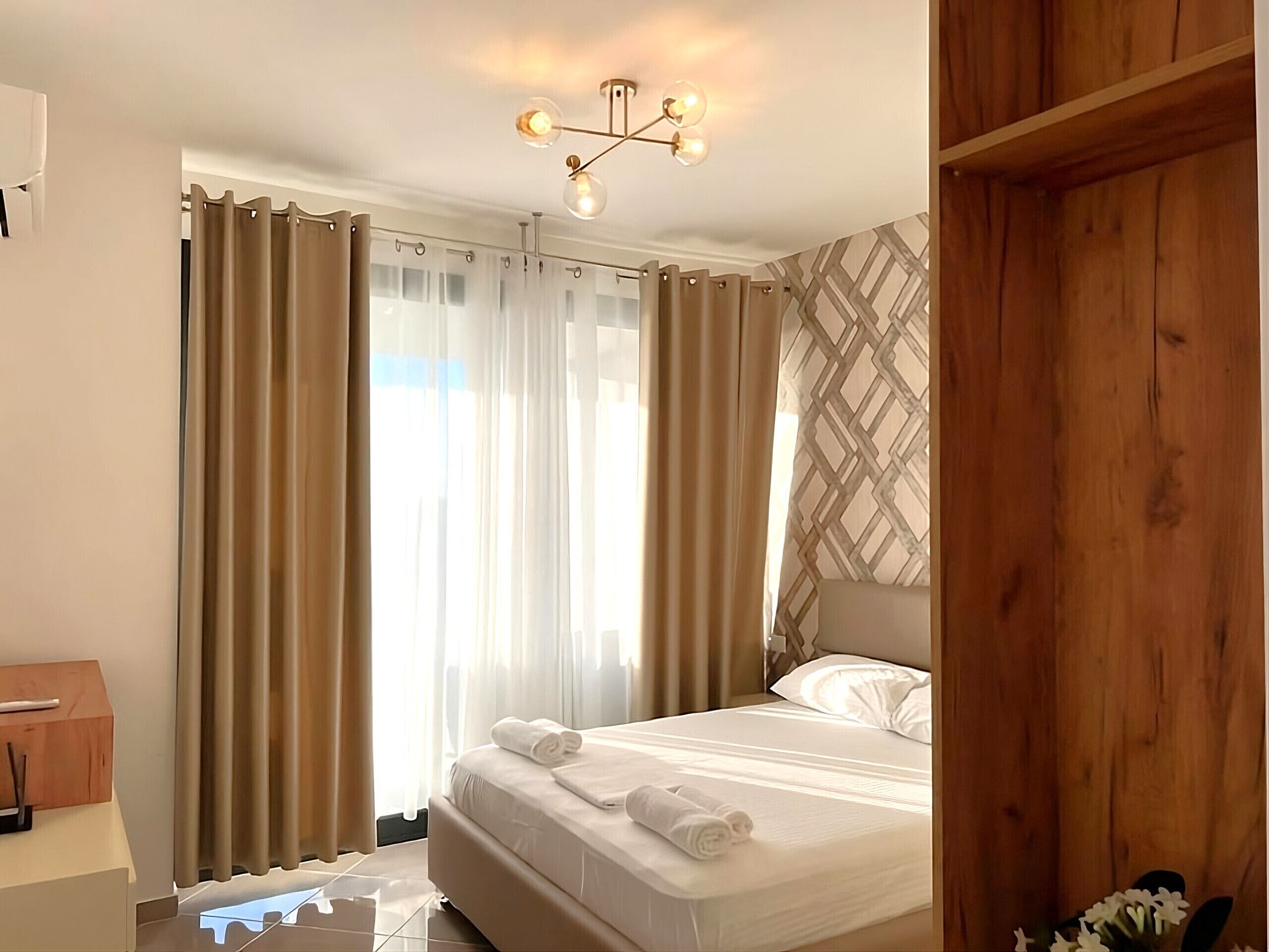 A hotel room with a bed, curtains, a wooden wardrobe, and a ceiling light fixture.