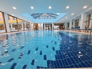 An indoor swimming pool with clear blue water, surrounded by lounge chairs and a glass ceiling.