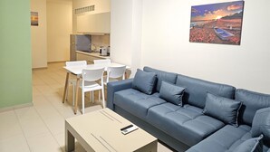 Een moderne woonkamer met een blauwe bank, een eettafel met stoelen, een keuken met een koelkast, en een schilderij van een strandlandschap aan de muur.