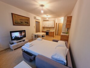 A hotel room with a bed, a TV, a desk, a chair, a small kitchenette, and a painting on the wall. - Teo's K Apartment - One Bedroom (Vlorë)