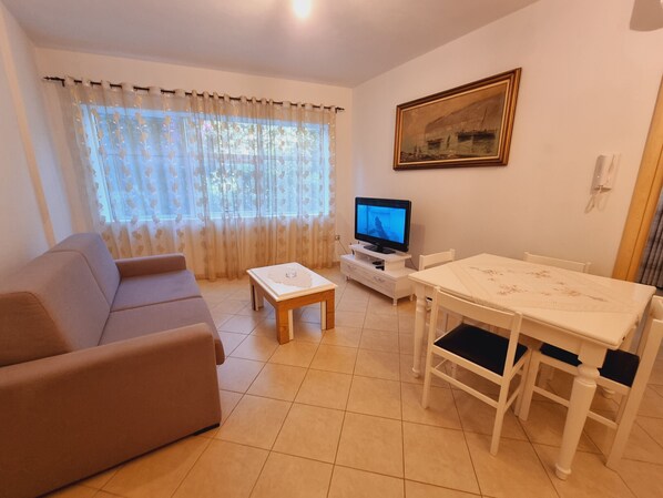A living room with a sofa, a coffee table, a TV on a stand, a dining table with chairs, and a painting on the wall. - Teo's K Apartment - One Bedroom (Vlorë)