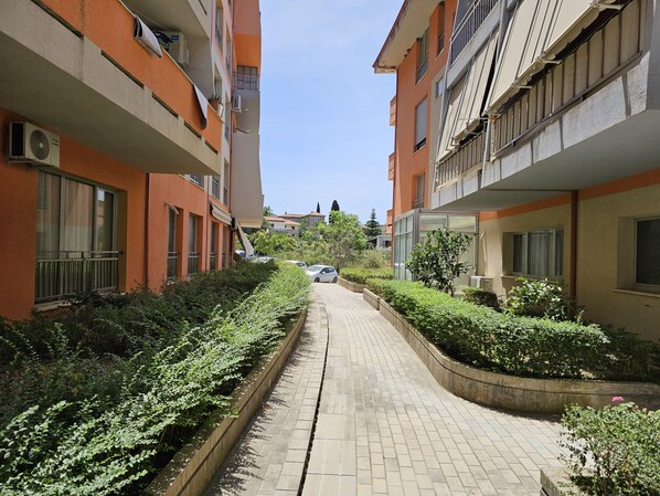 A narrow pathway flanked by multi-story buildings with balconies, air conditioning units, and a clear sky.