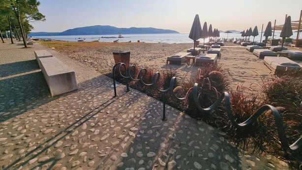 A beachside walkway with sun loungers and umbrellas, a trash bin, and a clear view of the sea and mountains in the background. - Teo's Deluxe Studios - Balcony Triple Studio (Vlorë)
