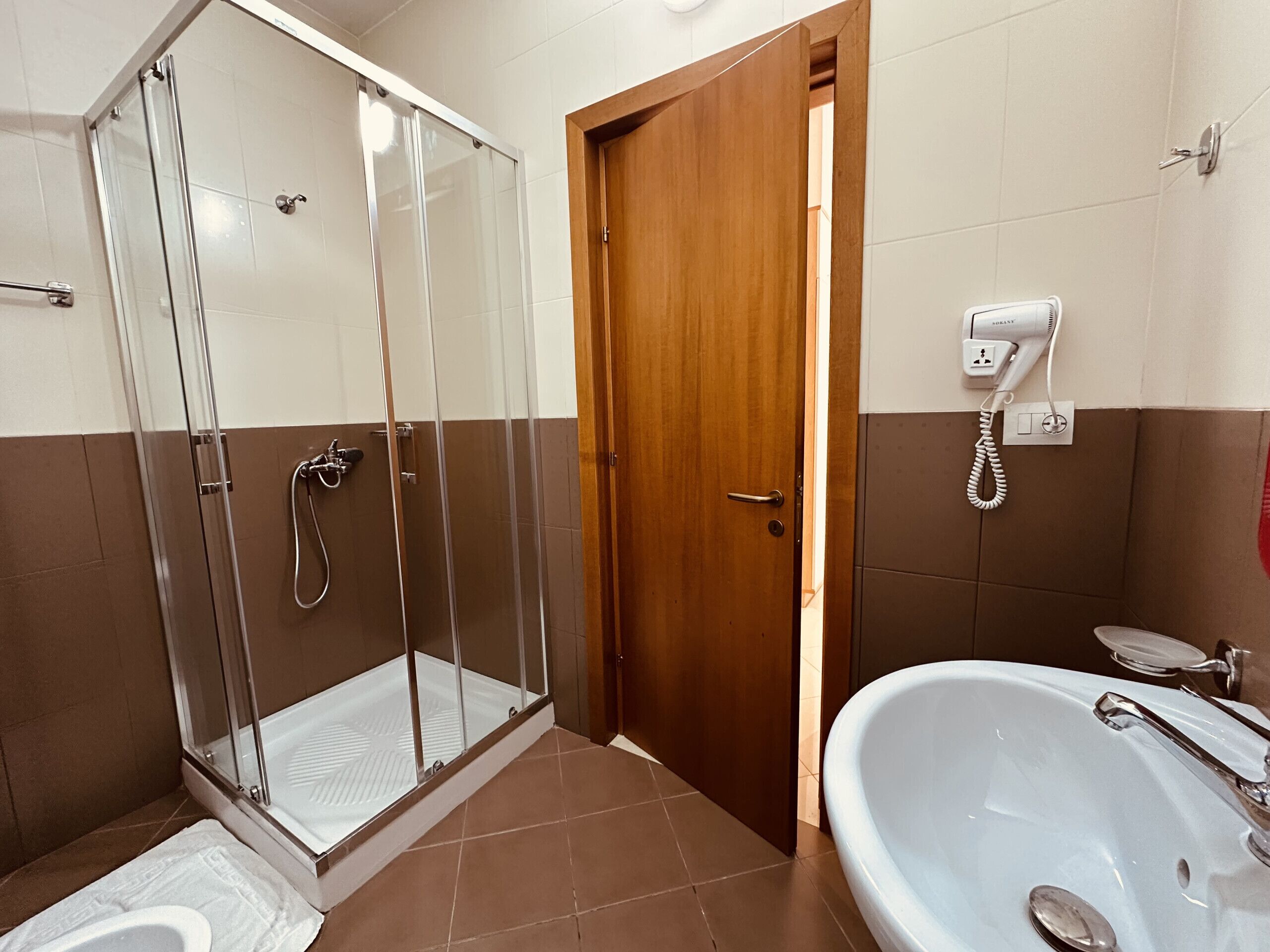 A bathroom with a glass-enclosed shower, a wooden door, a white sink with a mirror, and a hairdryer on the wall.
