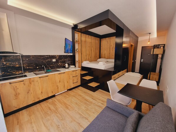 A compact hotel room with a kitchenette, a bed, a sofa, and a small dining table.