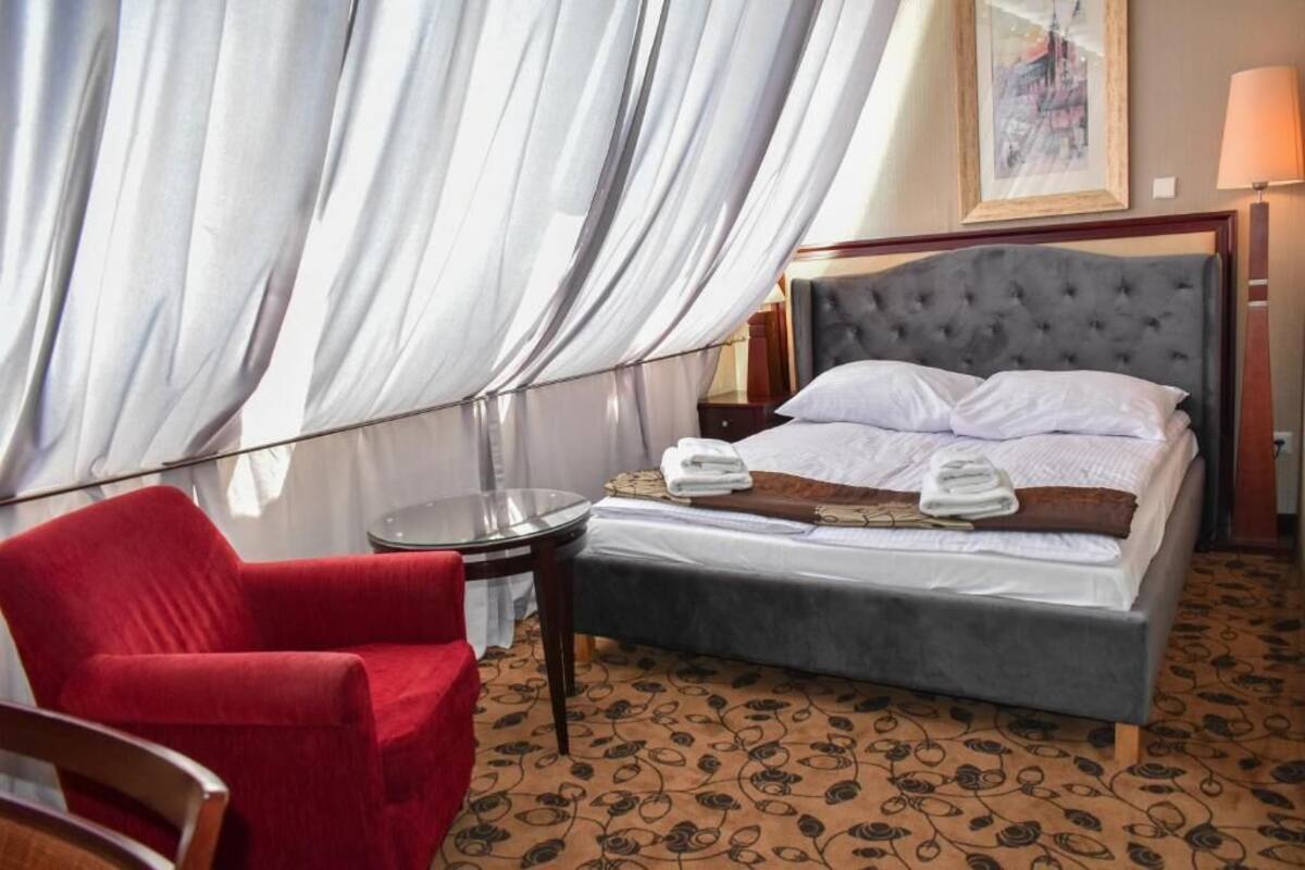 A hotel room with a bed, a red armchair, a small table, and a window with sheer curtains.