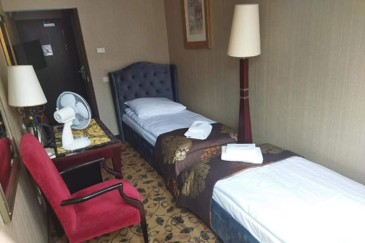 A hotel room with two beds, a red chair, a desk with a lamp, a mirror, and a standing fan.