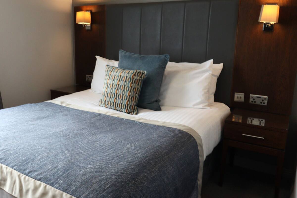 Un lit bien fait, avec une couverture bleue et des coussins à motifs, une tête de lit en bois et des lampes de chevet.