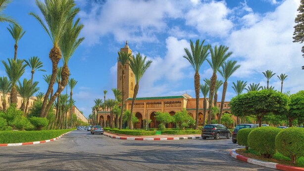 A street leading to a building with a prominent tower, surrounded by palm trees and parked cars. - Riad Amirkesh and Pool (Marrakech)