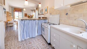 A kitchen with a gas stove, white cabinets, a sink, and a dining area with a table and chairs. - Appartamenti Costa Sud Gallipoli (Torre Suda)