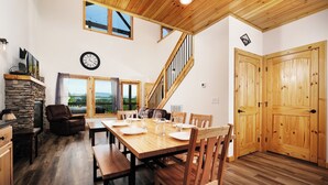 A spacious dining area with a wooden table set for four, a stone fireplace, and a view of the outdoors through large windows. - Smokey's Hideaway: Your Secret in the Smokies (Sevierville)
