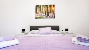 Ein ordentlich bezogenes Bett mit einer violetten Decke, zwei Nachttische mit Lampen und ein Waldgemälde an der Wand.