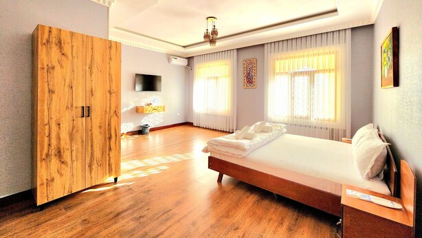 A hotel room with a wooden wardrobe, a bed with white linens, a flat-screen TV mounted on the wall, and large windows with sheer curtains. - Ancient Tagayev Hotel (Bukhara)