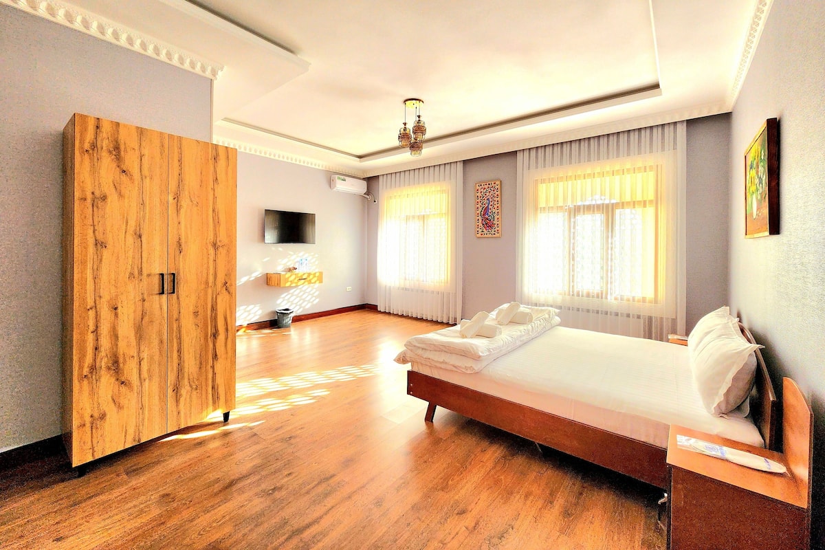 Habitación de hotel con un armario de madera, una cama con ropa blanca, un televisor de pantalla plana montado en la pared y ventanas grandes con cortinas translúcidas.
