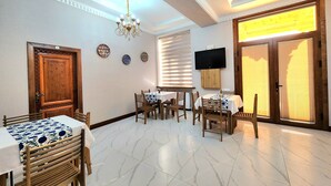 A dining area with a chandelier, wooden chairs, a table with a patterned tablecloth, and a television mounted on the wall. - Ancient Tagayev Hotel (Bukhara)