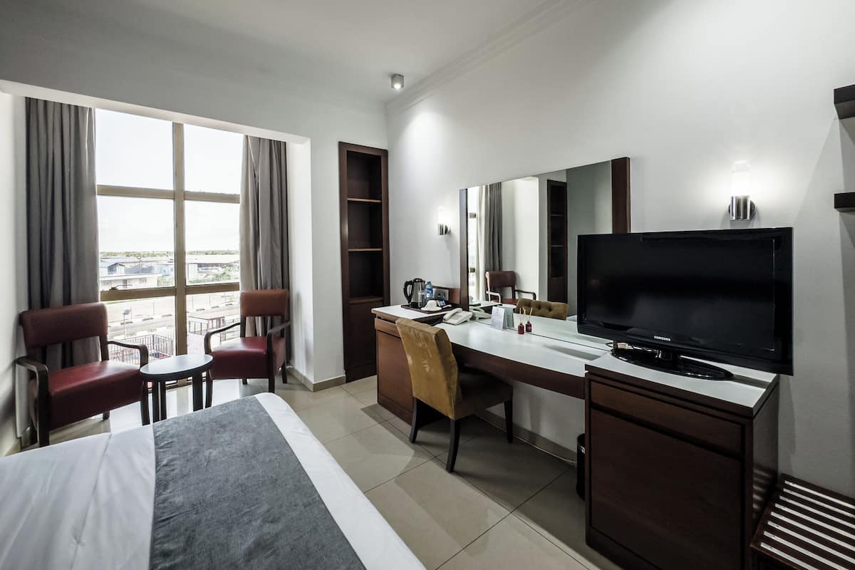 A hotel room with a bed, a desk, a chair, a television, and a large window offering a view of the city.