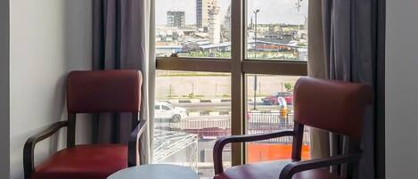 A room with two red chairs and a small table, large windows with curtains, and a view of a parking lot and buildings outside.