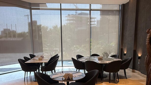 A modern dining area with round tables and black chairs, large windows offering a view of a body of water and buildings, and a decorative wall sculpture. - Avenue Suites (Lagos)