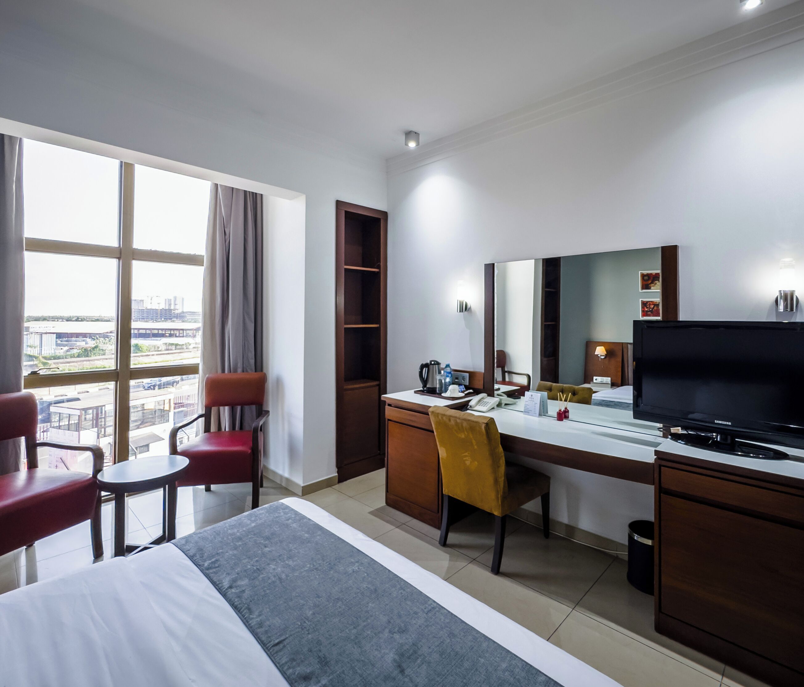A hotel room with a bed, a desk with a computer, a chair, a television, and a window with a view of buildings.