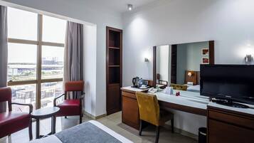A hotel room with a bed, a desk with a computer, a chair, a television, and a window with a view of buildings.