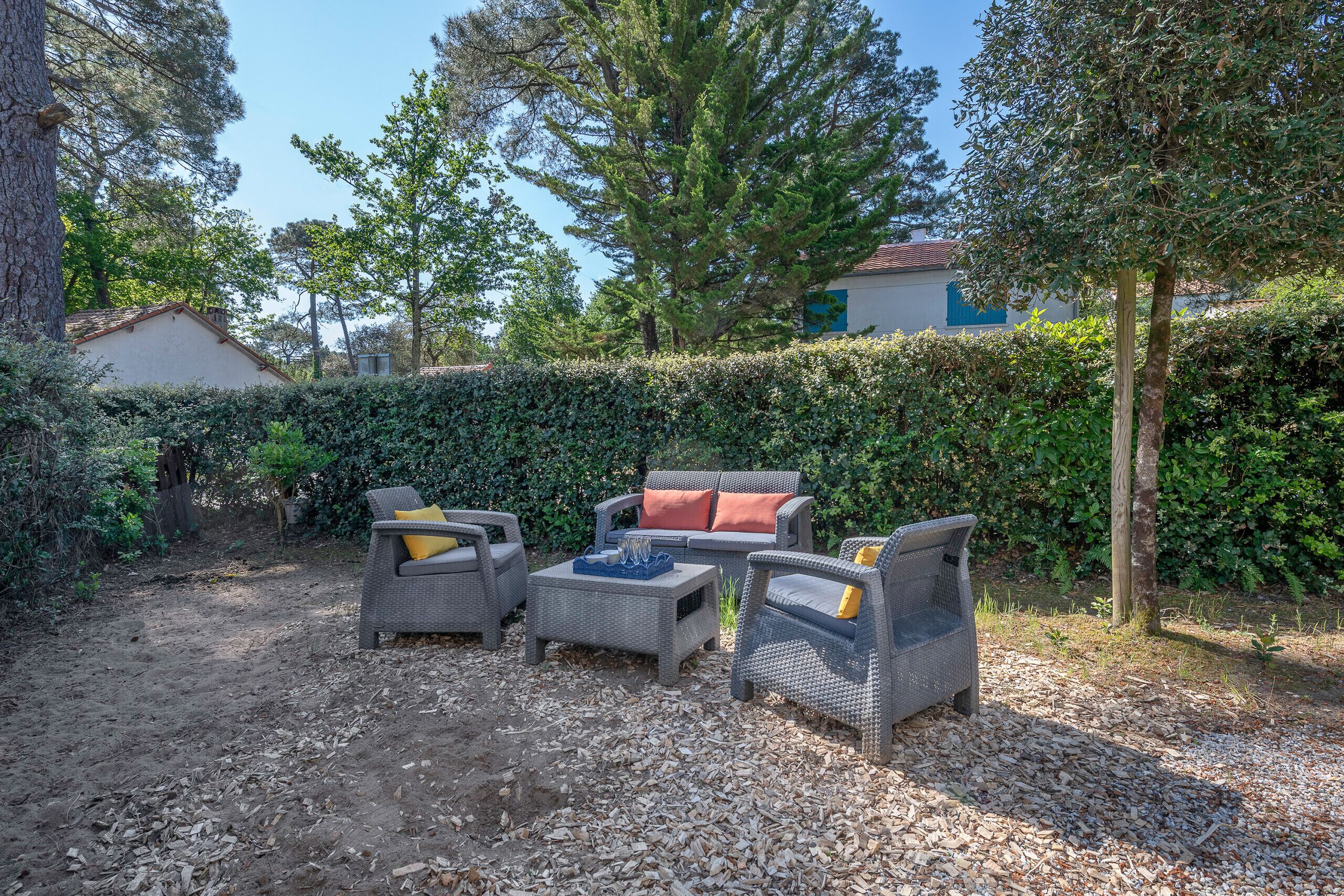 Un espace extérieur aménagé avec des meubles en osier et une petite table, entouré d’une haie et d’arbres.