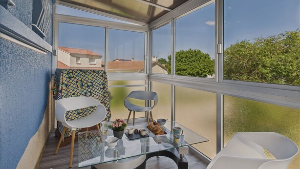 Una veranda moderna con ampie finestre, un tavolo in vetro, due sedie e vista sugli edifici circostanti.
