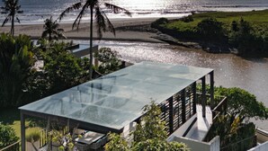 A modern glass-roofed structure with outdoor seating, overlooking a beach and water.