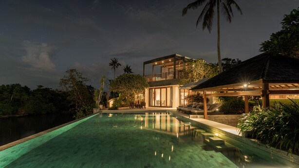 A nighttime view of a modern resort with a swimming pool, a covered outdoor area, and lush tropical vegetation.
